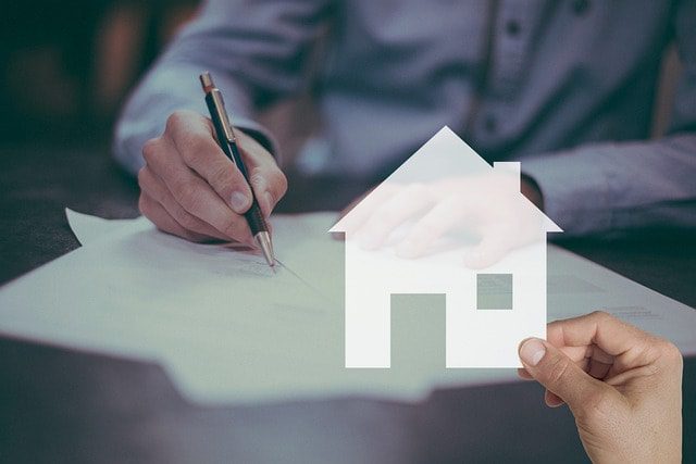 Person signing financial documents with a house icon overlay, symbolizing home loan refinancing and loan-to-value ratio calculations.