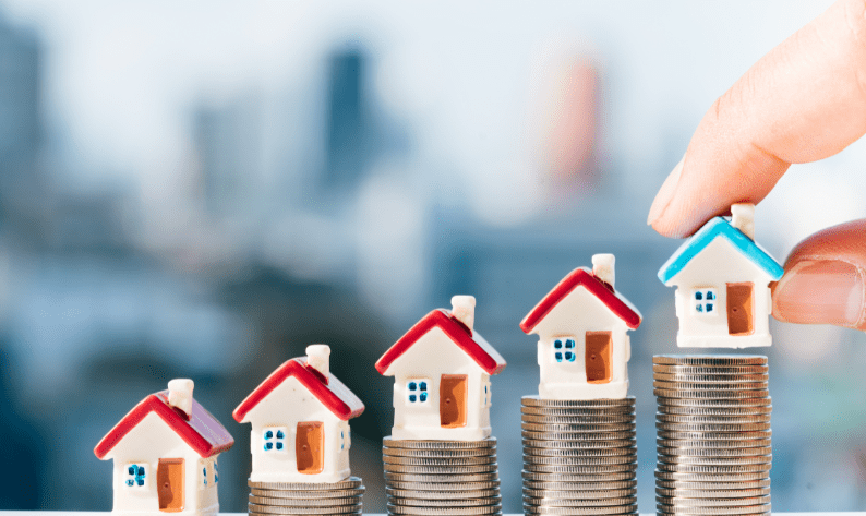 Miniature houses placed on ascending stacks of coins, with a hand placing the highest-value house—symbolising growing property investment and wealth accumulation.