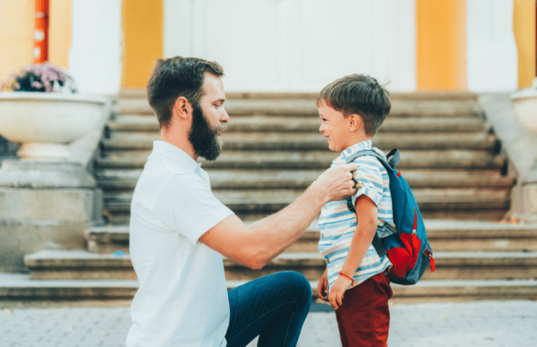 Father kneeling to help his son with a backpack in front of a school or building.