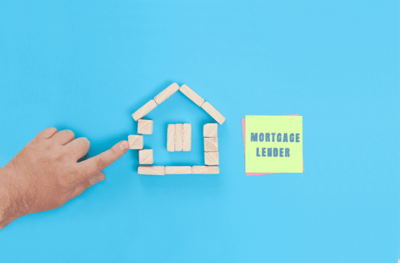 Hand pointing at a wooden block house beside a note reading “Mortgage Lender” on a blue background.