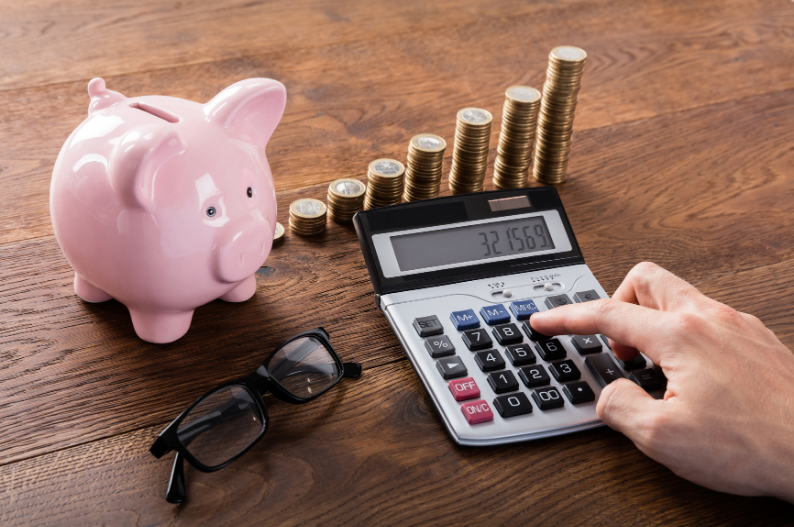 Person using a calculator beside a pink piggy bank and stacked gold coins, symbolising financial savings and planning.
