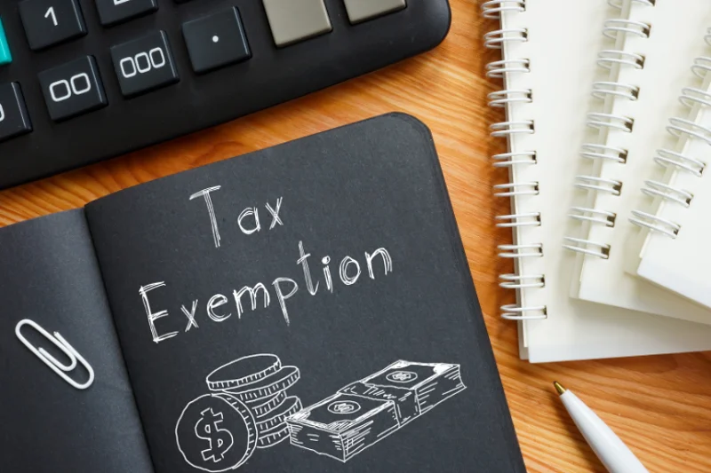 Notebook with the words "Tax Exemption" written on the cover, next to coins, cash illustrations, a pen, calculator, and notepads on a wooden desk—symbolising financial planning and tax relief.