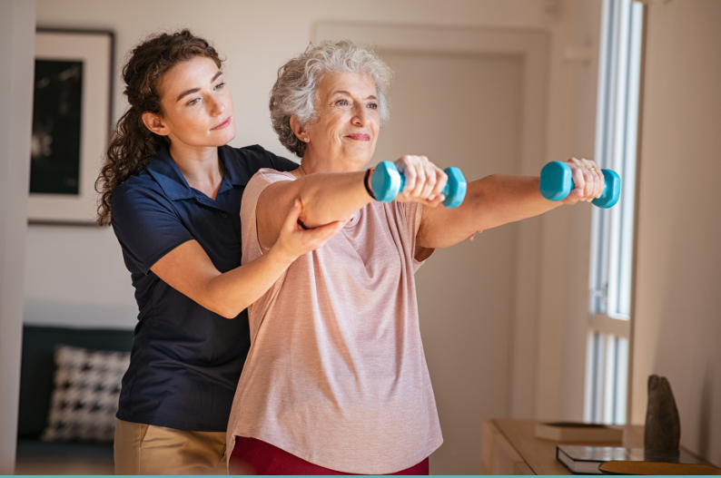 Physiotherapist assisting an elderly woman with arm exercises at home using small dumbbells.
