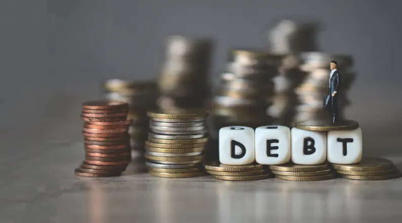 Miniature man standing on stacked coins next to dice spelling “DEBT”, symbolising the burden and management of debt.