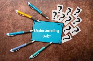 Pens pointing toward a blue card labeled “Understanding Debt,” surrounded by question marks on a brown surface.
