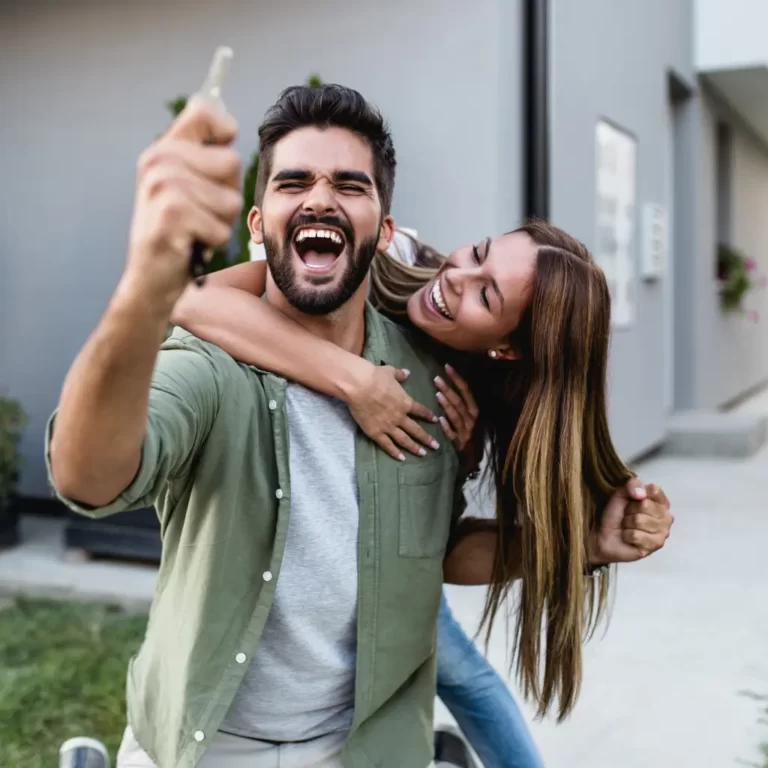Excited young couple celebrating new home purchase while holding house keys outside modern property.