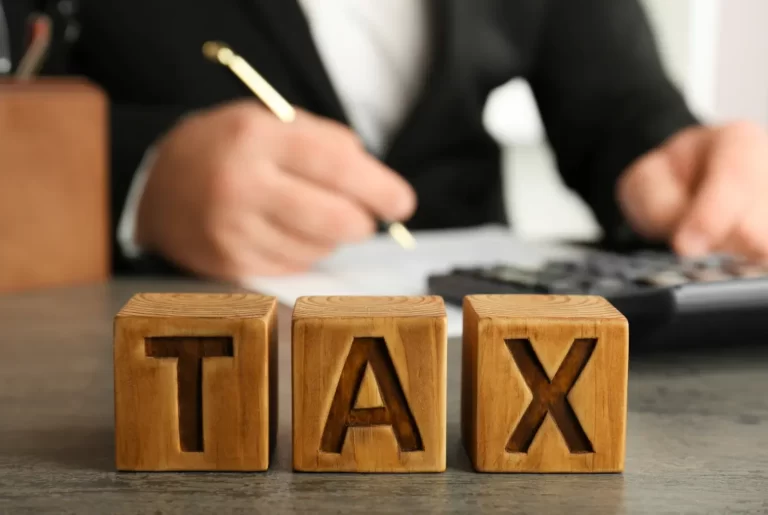 Wooden blocks spelling “TAX” with person calculating