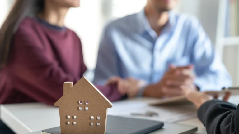Close-up of a wooden house model on a desk while a couple meets with a mortgage broker to discuss NSW First Home Buyer Grant and home loan eligibility.