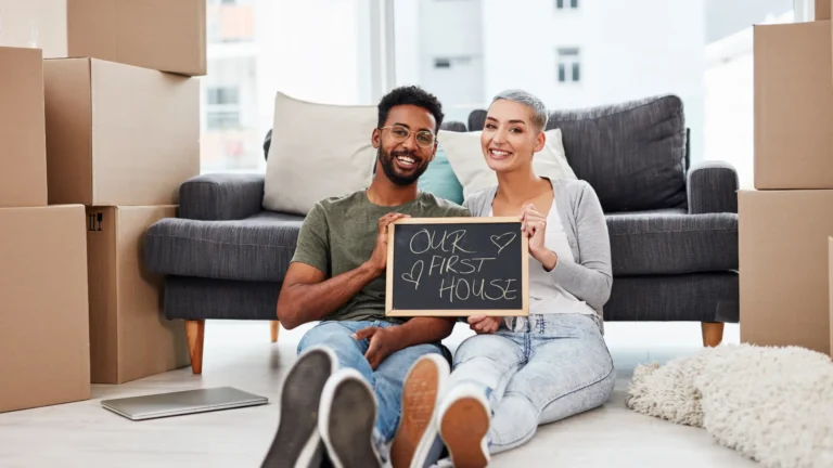 Smiling couple holding a chalkboard that says “Our First House,” surrounded by moving boxes in their new Australian home, symbolising first home ownership success.